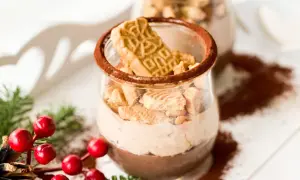 Zwei Gläser mit cremiger Spekulatius-Creme, garniert mit Spekulatius-Stücken und Kakaopulver. Weihnachtliche Deko mit Tannenzweigen und roten Beeren.