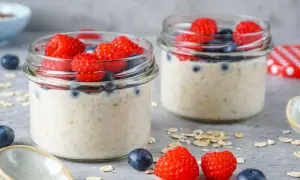 Zwei Gläser mit Overnight Oats, garniert mit frischen Himbeeren und Blaubeeren. Ein Löffel und Haferflocken liegen daneben.