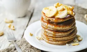 Ein Stapel goldbrauner Bananenpfannkuchen, beträufelt mit Sirup und garniert mit Bananenscheiben und Cashewkernen.