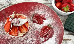 Zuckersüße Erdbeer Tartelettes, bestäubt mit Puderzucker, auf rotem Teller. Daneben frische Erdbeeren und eine kleine Schale voller roter Früchte.