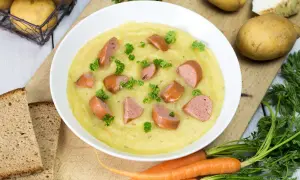 Cremige Kartoffelsuppe mit Würstchenstücken und Petersilie, zubereitet im Thermomix. Dazu gibt es Brot und frische Karotten.