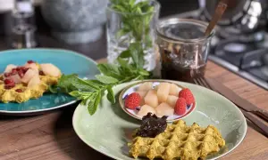 Zwei Teller mit goldgelben Haferflocken Waffeln, garniert mit Früchten und Marmelade, daneben frische Minze.