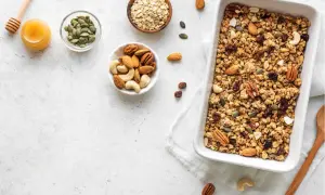 Zutaten für ein leckeres baked oats rezept: Haferflocken, Nüsse, Kürbiskerne, Rosinen und Honig in Schalen, daneben eine Auflaufform mit fertigem Granola.