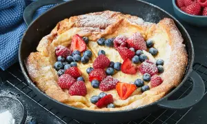 Ein goldbrauner Dutch Baby mit Puderzucker und frischen Beeren wie Erdbeeren, Himbeeren und Blaubeeren in einer Gusseisenpfanne.