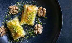 Zwei goldbraune Baklava-Stücke, bestreut mit gehackten Pistazien und Walnüssen, auf einem dunklen Teller. Ein köstliches Baklava Rezept.