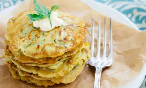 Ein Stapel goldbrauner Zucchini-Pancakes, garniert mit einem Klecks Sour Cream und Petersilie, daneben eine Gabel.