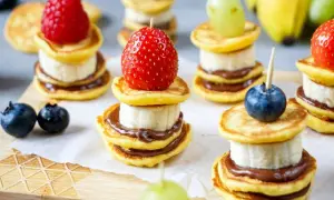 Kleine, gestapelte Baby-Pfannkuchen mit Banane und Schokoaufstrich, garniert mit Beeren und Trauben. Ein süßer Genuss!