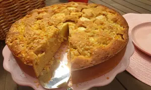 Ein goldbrauner Apfelkuchen ohne Zucker, bestreut mit Nüssen, wird auf einem rosa Teller präsentiert. Daneben liegt ein roter Apfel.