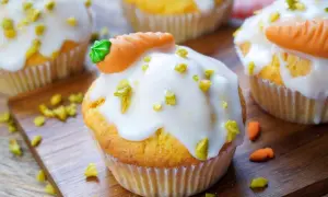 Muffins mit weißer Glasur, gehackten Pistazien und Marzipan-Karotten auf Holzbrett.