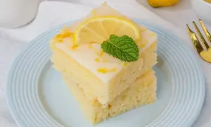 Zwei Stücke zitronenkuchen mit joghurt, garniert mit Zitronenscheibe und Minze, auf blau-weiß gestreiftem Teller.