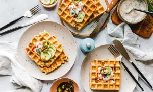 Drei Teller mit veganen Waffeln, garniert mit Gurkenscheiben, Zwiebeln und einer cremigen Sauce. Dazu ein Schälchen mit eingelegten Jalapeños.