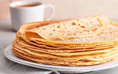 Ein Stapel goldbrauner Pfannkuchen, bestäubt mit Puderzucker, wartet auf ein leckeres Pfannkuchen Rezept. Daneben eine Tasse Kaffee.