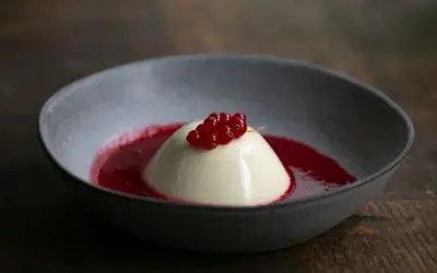 Cremige Panna Cotta mit roter Beerensauce und frischen Johannisbeeren in einer grauen Schale auf Holzhintergrund.