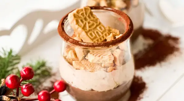 Zwei Gläser mit cremiger Spekulatius-Creme, garniert mit Spekulatius-Stücken und Kakaopulver. Weihnachtliche Deko mit Tannenzweigen und roten Beeren.