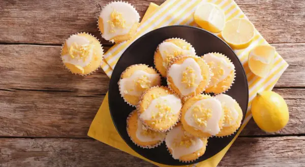 Zitronen-Muffins, frisch aus dem Thermomix, mit Zuckerguss und Zitronenschale bestreut, auf einem Holztisch.