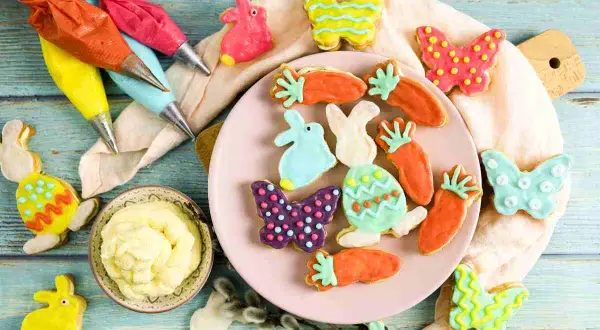 Bunte Osterkekse in Hasen-, Karotten- und Schmetterlingsformen, bereit für ein einfaches Osterkekse Rezept. Spritzbeutel mit Zuckerguss liegen bereit.
