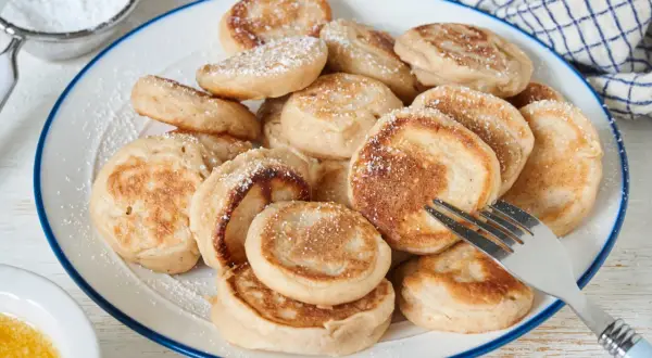Ein Teller voller goldbrauner Poffertjes, bestäubt mit Puderzucker. Ein Gabel liegt bereit, um das leckere Poffertjes Rezept zu genießen.