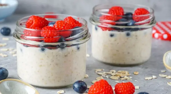 Zwei Gläser mit Overnight Oats, garniert mit frischen Himbeeren und Blaubeeren. Ein Löffel und Haferflocken liegen daneben.