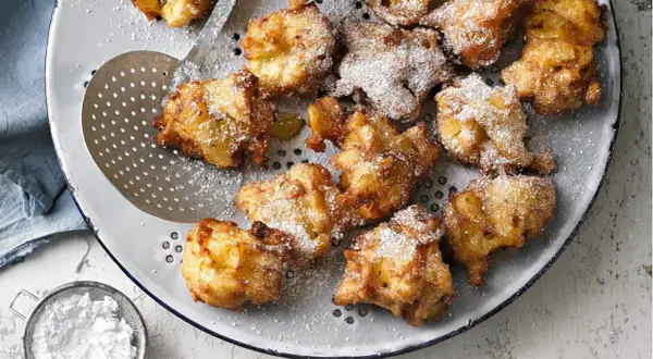 Goldbraune Apfelkrapfen, frisch aus der Pfanne, mit Puderzucker bestäubt, liegen auf einem Sieb.
