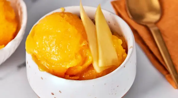 Erfrischendes Mango-Sorbet in einer Schale, garniert mit Mangostücken. Daneben liegt ein goldener Löffel auf einem orangen Tuch.