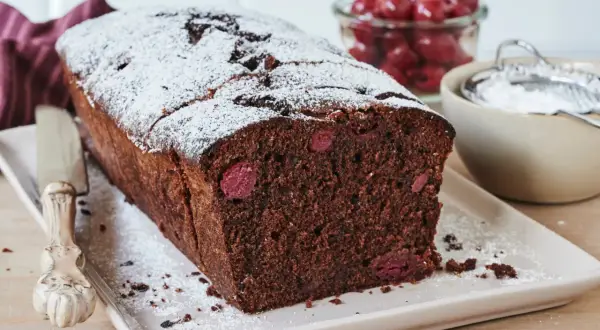 Saftiger schoko kirsch kuchen, bestäubt mit Puderzucker. Daneben eine Schale mit Kirschen und ein Sieb.