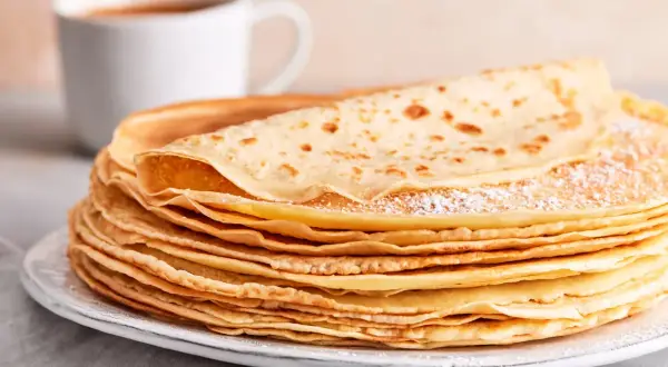 Ein Stapel goldbrauner Pfannkuchen, bestäubt mit Puderzucker, wartet auf ein leckeres Pfannkuchen Rezept. Daneben eine Tasse Kaffee.