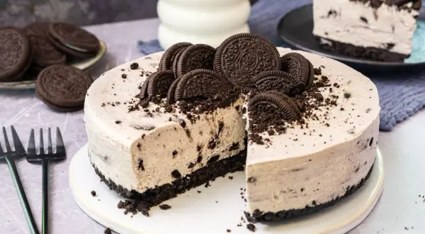 Ein köstlicher Oreo Kuchen, dekoriert mit ganzen und zerbrochenen Keksen, wartet darauf, angeschnitten zu werden. Daneben liegen Gabeln und ein Glas Milch.