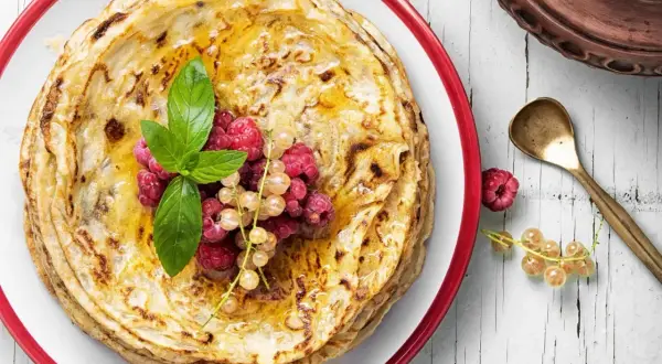 Ein Stapel goldbrauner Pfannkuchen, garniert mit Beeren und Minze. Der Pfannkuchenteig ist perfekt gebacken und mit Honig beträufelt.