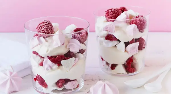 Zwei Gläser mit Himbeer Baiser Dessert, geschichtet mit Sahne, frischen Himbeeren und Baiserstückchen.