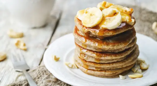 Ein Stapel goldbrauner Bananenpfannkuchen, beträufelt mit Sirup und garniert mit Bananenscheiben und Cashewkernen.