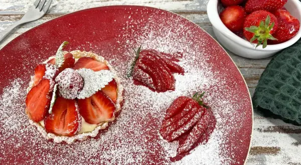 Zuckersüße Erdbeer Tartelettes, bestäubt mit Puderzucker, auf rotem Teller. Daneben frische Erdbeeren und eine kleine Schale voller roter Früchte.