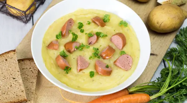 Cremige Kartoffelsuppe mit Würstchenstücken und Petersilie, zubereitet im Thermomix. Dazu gibt es Brot und frische Karotten.