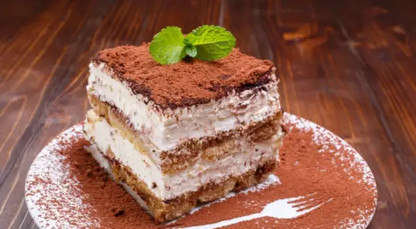 Ein Stück cremiges Tiramisu ohne Ei, bestäubt mit Kakao und garniert mit Minze, auf einem Teller mit Kakaopulver-Gabelabdruck.