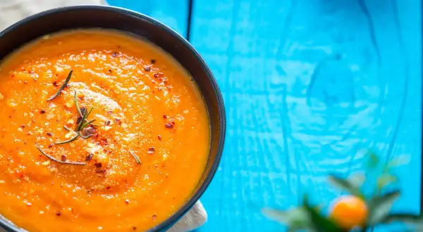 Eine cremige Tomatensuppe, zubereitet im Thermomix, garniert mit Rosmarin und Chiliflocken, serviert auf einem blauen Holztisch.