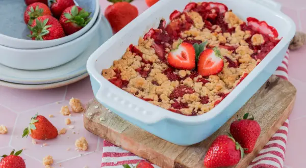 Erdbeer-Crumble im Auflaufform, garniert mit frischen Erdbeeren, daneben eine Schüssel mit Erdbeeren.