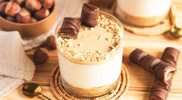 Ein Glas mit cremiger Kinder Bueno Creme, garniert mit Nüssen und Schokostückchen. Daneben liegen ganze Kinder Bueno Riegel.