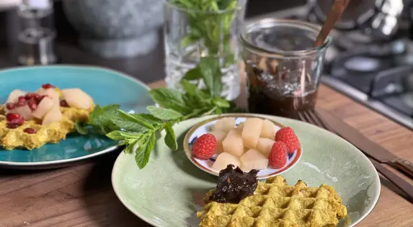 Zwei Teller mit goldgelben Haferflocken Waffeln, garniert mit Früchten und Marmelade, daneben frische Minze.