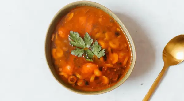 Herzhaftes Soljanka Rezept: Eine Schüssel voller würziger Suppe mit Oliven, Kapern und Kräutern, daneben ein goldener Löffel.