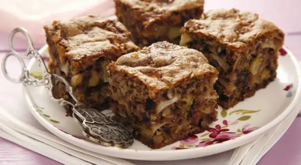 Vier Stücke saftiger Apfel Walnuss Kuchen auf einem Teller mit floralem Muster, daneben eine antike Kuchenzange.