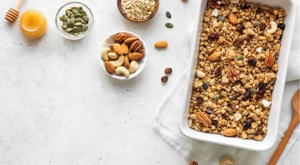 Zutaten für ein leckeres baked oats rezept: Haferflocken, Nüsse, Kürbiskerne, Rosinen und Honig in Schalen, daneben eine Auflaufform mit fertigem Granola.