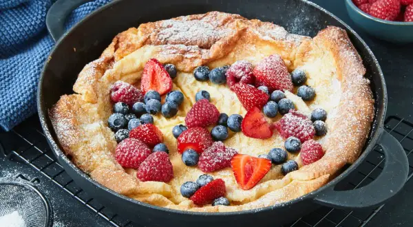 Ein goldbrauner Dutch Baby mit Puderzucker und frischen Beeren wie Erdbeeren, Himbeeren und Blaubeeren in einer Gusseisenpfanne.