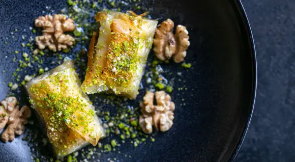 Zwei goldbraune Baklava-Stücke, bestreut mit gehackten Pistazien und Walnüssen, auf einem dunklen Teller. Ein köstliches Baklava Rezept.