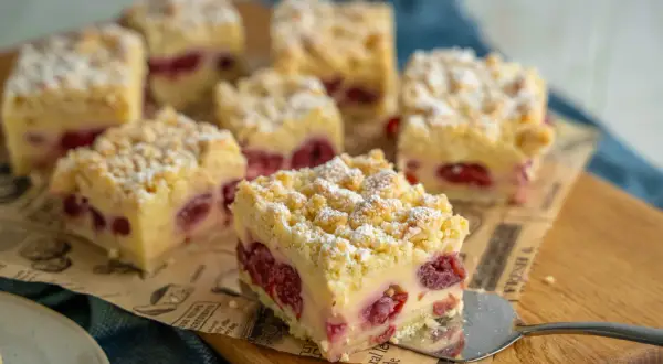Saftiger Kirschkuchen mit Pudding und Streuseln, bestäubt mit Puderzucker. Ein Stück wird gerade serviert.