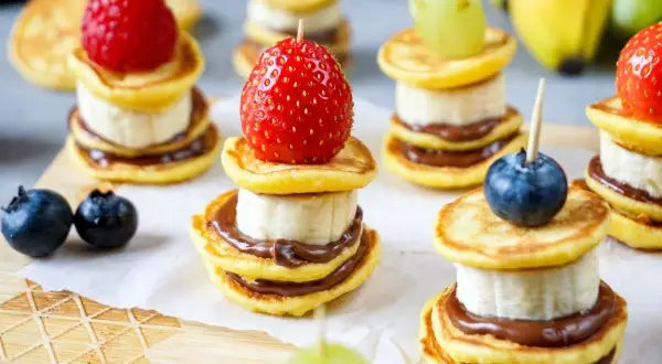 Kleine, gestapelte Baby-Pfannkuchen mit Banane und Schokoaufstrich, garniert mit Beeren und Trauben. Ein süßer Genuss!