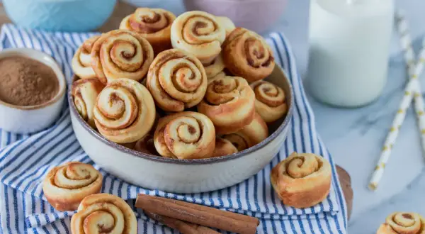 Mini Zimtschnecken in einer Schüssel, daneben Zimtstangen und ein Glas Milch.