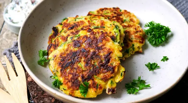 Goldbraune zucchini puffer, garniert mit Petersilie, liegen auf einem Teller. Daneben eine Schüssel mit Dip.