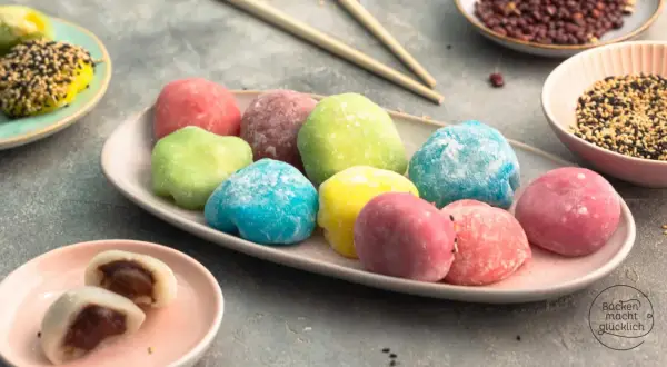 Bunte Mochi-Bällchen, die Lust aufs **Mochis selber machen** machen. Auf einem Teller liegen sie in verschiedenen Farben, daneben Schüsseln mit Sesam und roten Bohnen.