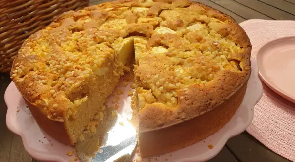 Ein goldbrauner Apfelkuchen ohne Zucker, bestreut mit Nüssen, wird auf einem rosa Teller präsentiert. Daneben liegt ein roter Apfel.