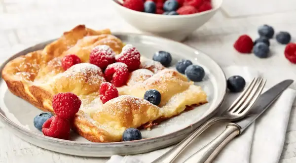 Ein goldbrauner Ofenpfannkuchen, bestäubt mit Puderzucker und garniert mit frischen Himbeeren und Blaubeeren. Perfekt für ein leckeres Ofenpfannkuchen Rezept.