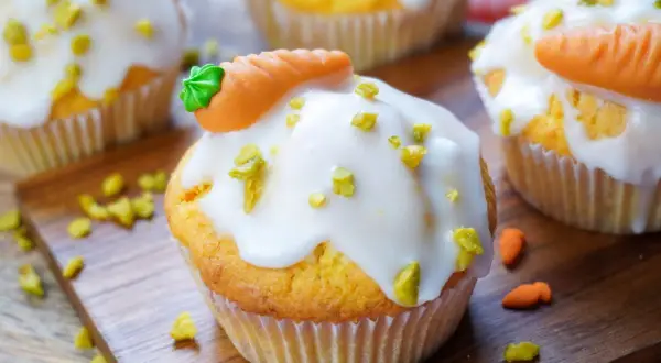 Muffins mit weißer Glasur, gehackten Pistazien und Marzipan-Karotten auf Holzbrett.